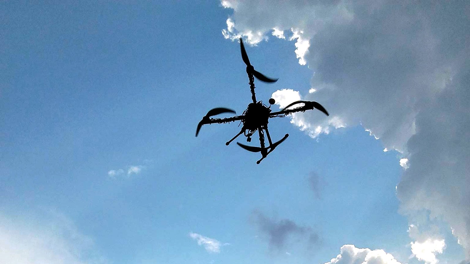 Quadcopter silhouette flying against a bright cloudy sky.